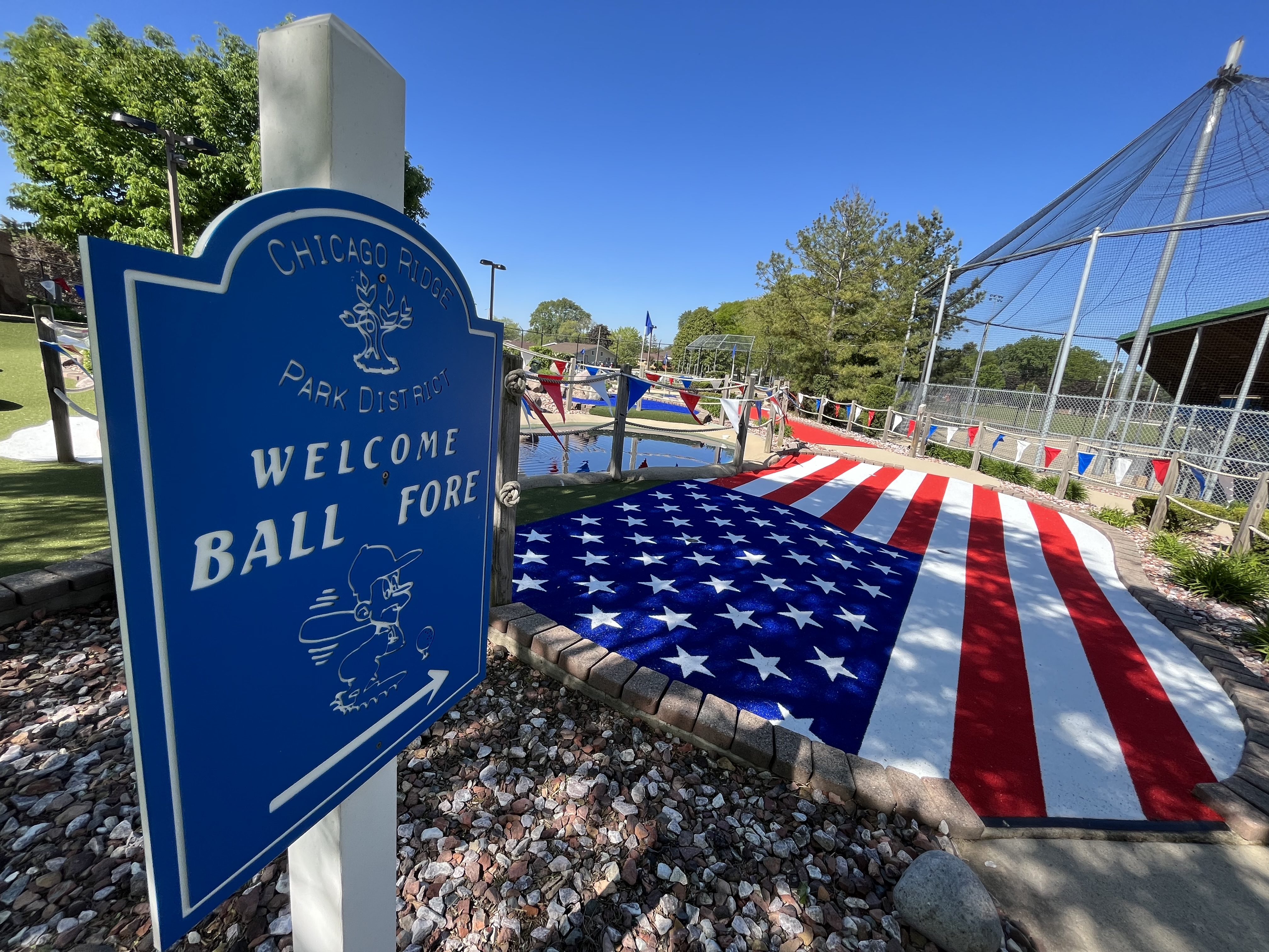 a blue sign wtih welcome to ball fore on it in white text and an american flag in the ground to the right 